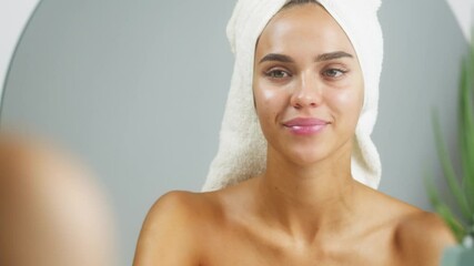 Woman using moisturizing lipstick and smiling, morning skincare routine. Young female student with bare shoulders wearing towel on head looking in mirror in spa salon. Natural day make up concept. - Powered by Adobe