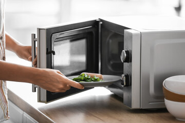 Young woman putting plate with food into microwave oven in kitchen, closeup