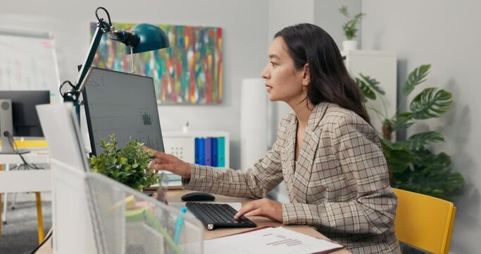 An attractive, experienced company accountant sits in office in front of a computer checking statistical data employee salary charts comparing paper diagrams with electronic ones financial analyst