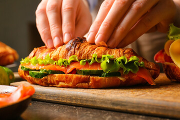 Woman preparing tasty croissant sandwich at kitchen table