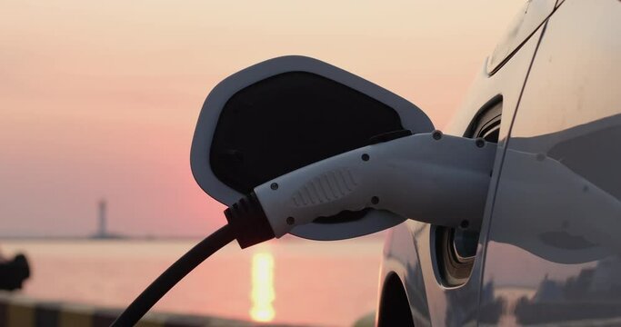 A man's hand pulls out the charging plug of an electric car from the car charging socket against the backdrop of the rising sun. Dawn of the era of electric transport.