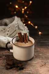 A mug of hot chocolate or cocoa with cinnamon sticks on a wooden table. Warm scarf and cozy autumn  winter concept. Blurred bokeh in the background.