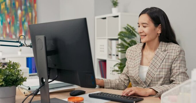 Woman with long brown hair wearing checkered jacket sits at desk in office in front of computer screen, receives call through webcam video chat waves to co-worker, shows documents she needs to correct