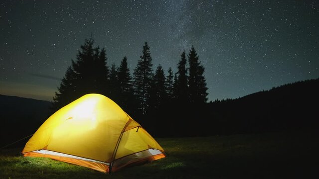 Time lapse of brightly illuminated camping tent glowing on campsite in dark mountains under night stars covered sky. Active lifestyle and trekking concept.