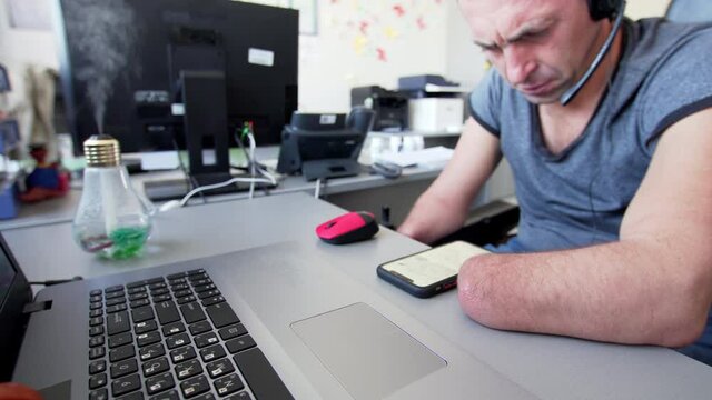 A serious caucasian man with a disability with amputated two stump hands and legs is reading something on his smartphone in the office. Focus is on the laptop at the desk in front of him