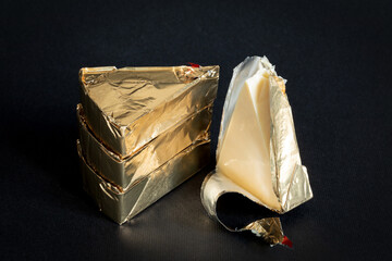 Thre triangular pieces of melted cream cheese wrapped in a golden aluminium foil and without packaging on a black background. Portioned triangular cheeses.