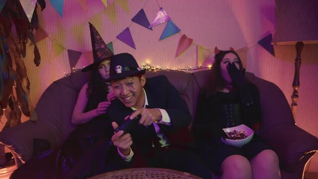 A guy in a creepy costume is actively playing a video game console, and the girls are sitting and bored at a Halloween party.