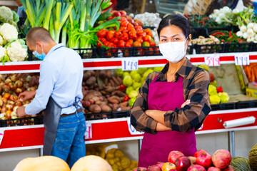 Oriental woman in uniform and face mask standing among shelves with fruits and vegetables and looking in camera. Her colleague working in background.