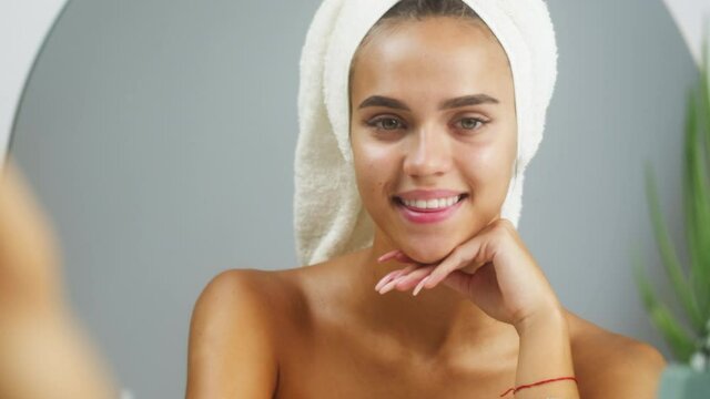 Happy woman smiling, looking in mirror in bathroom or spa. Portrait of young female student with bare shoulders and white towel on head after washing. Natural day make up, facial skin care concept.