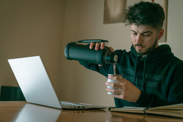 young man working / studying in front of a laptop while drinking a mate (Argentine drink)