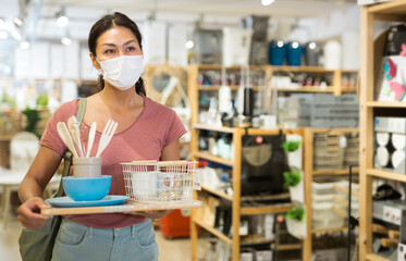 Asian woman in face mask walking through hall of housewares store and holding tray with tableware...