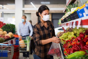 Oriental woman in face mask choosing red peppers in greengrocer. Man with cart shopping in background.