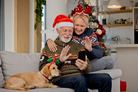 Senior elderly couple having video call over mobile phone celebrating Christmas at home with their yellow dog wearing Santa hats