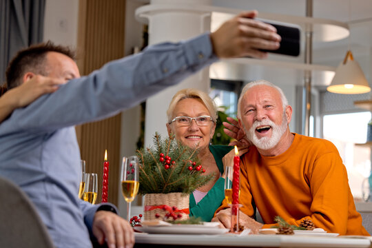 Happy family or friends sitting by Christmas decorated table and taking picture together or having video call using smart phone during festive season
