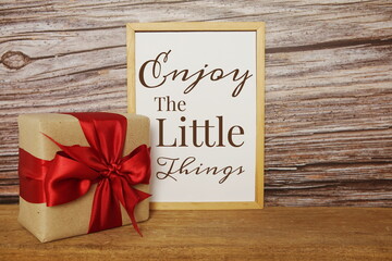 Blackboard with the text ENJOY THE LITTLE THINGS with gift box and red ribbon on wooden background