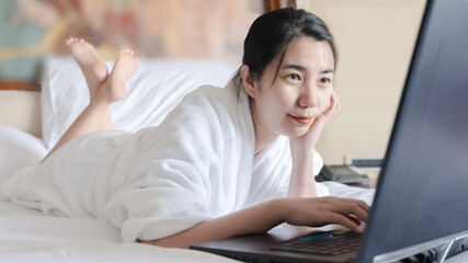 asian woman relaxing in the hotel and working with laptop on a bed after a shower with bathrobe. Freeland asia girl work on bed from hotel comfortably smiling enjoying care free time holiday