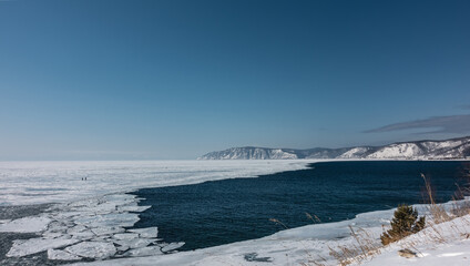 A non-freezing river flows out of a frozen lake. White ice floes on blue water. A mountain range against a blue sky. A sunny winter day. Baikal and Angara.