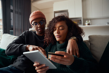 Multi-ethnic couple purchasing goods online on tablet using credit card, sitting on sofa in lounge