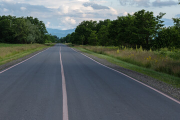 A suburban asphalt road with three solid lines of pink road markings. In the background of the mountain and the blue sky with clouds. The concept of travel.