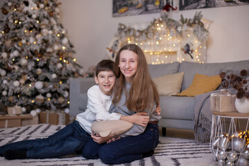 Happy brother and sister sitting on floor and fir tree with presents and looking at camera. Boy and girl at Christmas time. Merry Christmas and Happy New Year. Family values. Waiting for a holiday.