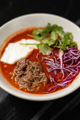 Mexican tomato soup, food with meat, parsley and sour cream in a white plate on a black background