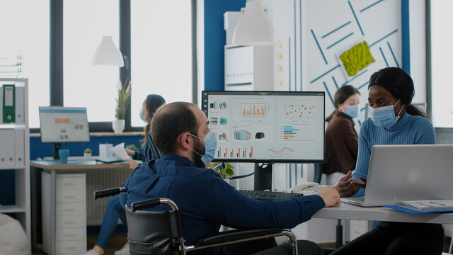 Diverse coworkers with protection face masks working together in new normal workplace during pandemic, invalid manager with disabilities pointing at laptop explaning to african woman company startegy