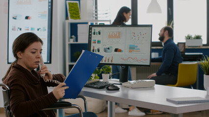 Busy entrepreneur with disabilities talking at phone with manager explaning financial statistics sitting in wheelchair in front of pc showing data, prosessing on display for project regarding economy