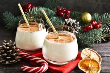 Two glass homemade winter eggnog with cinnamon, and fir tree branches, red napkin, cookies, sweet candy on wooden table. Traditional Christmas dessert