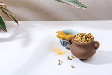 Cardamom in a ceramic pot and turmeric root on a white table.