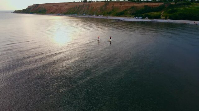 Men standing firmly on SUP board and paddling, pushing paddle through shining water and propelling paddle board at sunset near sea shore with car camping. Father and son actively spending time