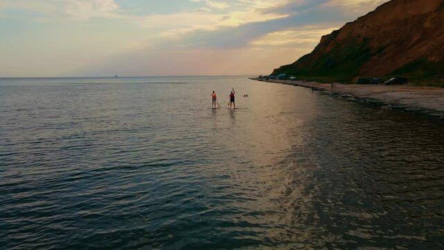 Men standing firmly on SUP board and paddling, pushing paddle through shining water and propelling paddle board at sunset near sea shore. Father and son actively spending time