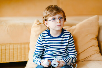Cute little blond kid boy with glasses playing with a video game console. Child having fun at home...