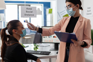 Businesswoman measuring temperature using infrared thermometer to prevent infection with coronavirus. Manager with medical face mask against covid19 working in business office during virus pandemic