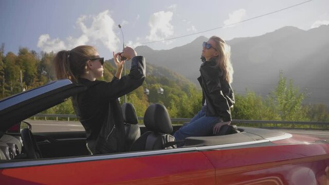 Beautiful girls take photos on a smartphone inside a red convertible. Funny photo shoot on vacation. Posing and enjoying the rest among the mountains.