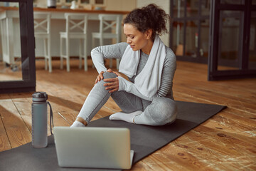 Female is sitting on mat and touching joint while suffering pain during video workout on laptop at...