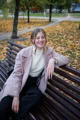 Stylish girl in a coat sits on a bench in the city and laughs