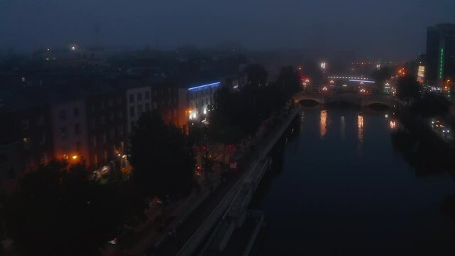 Forwards fly above Liffey river in misty evening. Aerial view of modestly illuminated waterfront. Dublin, Ireland