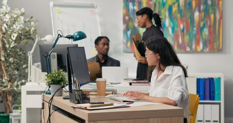Beautiful girl with glasses with distinctive Asian beauty dressed in white shirt, sits in office in front of computer, working, in background co-workers are discussing, talking, afternoon at company