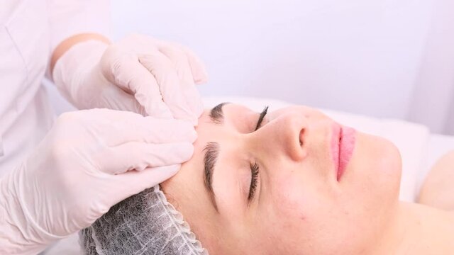 Young woman in a medical capon her head with closed eyes takes care of her skin face, receiving rejuvenate procedures by a beautician.