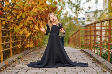 Fototapeta premium Happy laughing little girl on autumn trail in long magic black fashion dress. Halloween in gorgeous princess dress. Retro smile. Place for text. Laughing funny child girl in witch costume. road bridge
