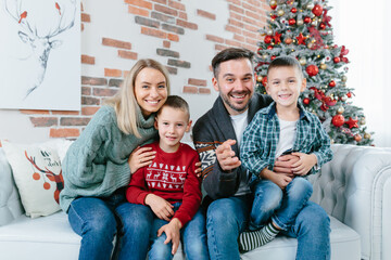 young family of four men woman husband and two little boys sons happily having fun together looking at webcam video communication with friends sitting on sofa at home near christmas and new year tree