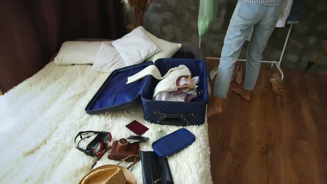 A belated girl puts clothes together with hangers in a suitcase standing on the bed, with accessories, against the background of an apartment with things