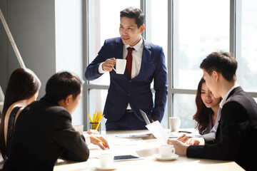 asian executive leader holding coffee cup standing to discussion with colleague in meeting on window office . businessman manager talking and advice to coworkers in conference . boss morning training