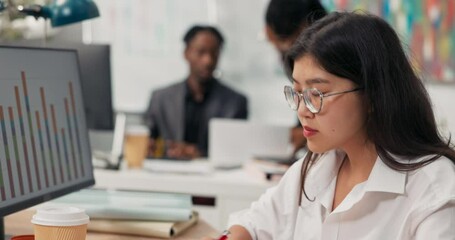 An attractive girl wearing glasses works at a desk in front of a computer, fills out documents, signs checks, in the background co-workers are browsing the project on the computer, talking, discussing