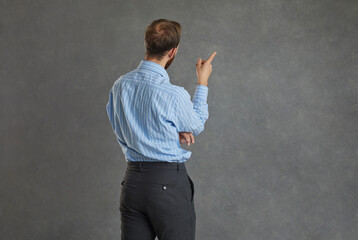 Focused businessman turned his back to the camera standing on a gray background. Man in a shirt ponders something pointing his finger at the free space for text. Planning and strategy concept. Banner.