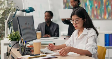 An attractive girl wearing glasses works at a desk in front of a computer, fills out documents, signs checks, in the background co-workers are browsing the project on the computer, talking, discussing