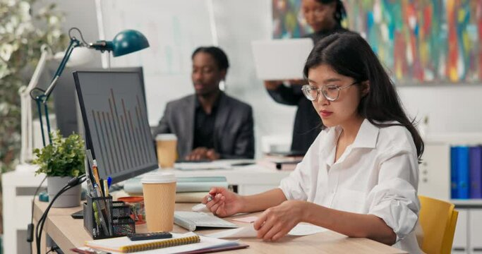 An attractive girl wearing glasses works at a desk in front of a computer, fills out documents, signs checks, in the background co-workers are browsing the project on the computer, talking, discussing