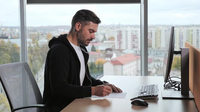 Serious man wearing casual clothes look ap pc screen writes ideas questions do review make research sit at desk. Businessman freelancer sitting in his workplace near panoramic window city background.