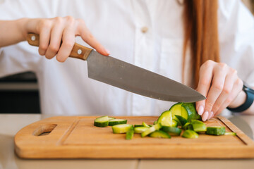 Close-up front view of unrecognizable woman cutting fresh cucumber cooking food salad sitting at wodden table in light kitchen room. Front view of female cooking vegetarian dieting salad at home.