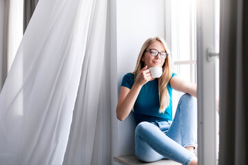 Young woman relaxing, smell and drink coffee on windowsill at home. Happy girl